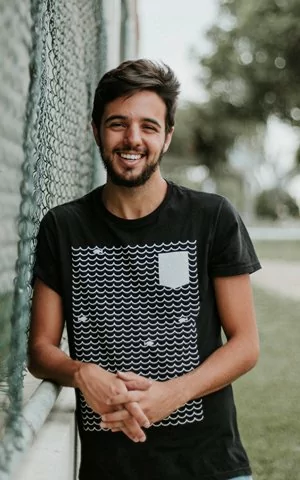 Man with a beard in a black shirt with white design leaning against a fence smiling