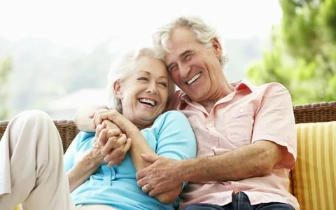 elderly couple sitting on yellow striped couch laughing together and holding hands 